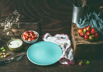 Empty blue plate on dark rustic wooden kitchen table with strawberries and yogurt in bowls. Country style food background with berries , still life. Place for your design, recipes , text or products