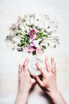 Female hands holding open jar of cream on white background with plants and flowers , top view. Natural cosmetic concept