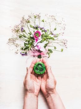Female hands holding green handmade soap on white background with plants and flowers, top view. Natural cosmetic concept