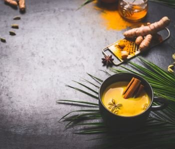 Black mug of vegan golden turmeric milk with spices on dark background with ingredients and palm leaves. Healthy hot drink. Immune boosting remedy , detox and dieting concept