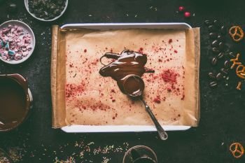 Spoon with melted chocolate on tray with red dried fruits powder topping on dark kitchen table background with various flavoring ingredients for chocolate bars making, top view. Clean food concept