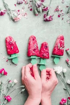 Female hands holding fruits popsicle ice. Homemade red ice cream on light green kitchen table background, top view. Healthy summer desserts. Frozen juices on sticks. Vegan ice. Summer food