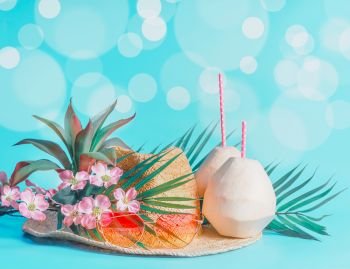 Female beach accessories with fresh coconut, drinking straws and tropical leaves and flowers , sunglasses and straw hat on sunny blue background with bokeh. Summer holiday concept, banner.
