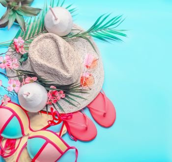Summer background with top view of woman’s summer beach accessories and coconut drinks: bikini,  flip flops, sunglasses, straw hat, palm leaves and tropical flowers on turquoise blue