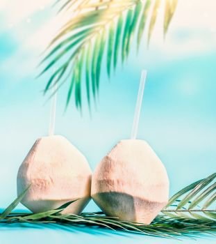 Two fresh coconut cocktails with tropical leaves at blue sky background with hanging palm leaves and sunshine. Tropical vacation. Summer holiday. Beach party. Healthy natural coconut water drink