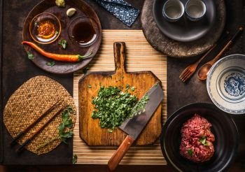 Chopped herbs on wooden cutting board with knife on dark rustic table background with kitchen utensils, bowls, plates and ingredients: Mincemeat, chili and soy sauce. Top view. Asian cuisine.