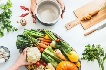Healthy lifestyle concept. Female hand prepare various colorful organic farm vegetables on white desk background with cooking pot. Vegetarian food and clean seasonal eating flat lay. Top view