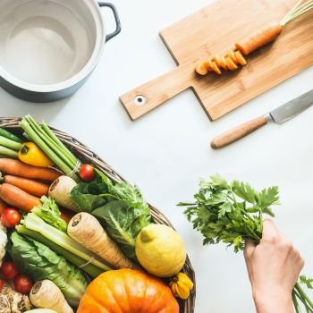Healthy seasonal vegetables with cooking pot , cutting board and knife on white desk background. Female hand holding fresh bunch of kitchen herbs. Clean eating. Farm organic vegetables. Top view