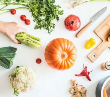 Female hand holding celery stalks on white table background  with pumpkin and various seasonal vegetables, cutting board and knife. Low carb cooking. Flat lay. Healthy lifestyle and eating. Dieting