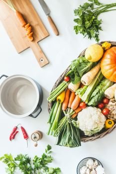 Variety of harvest organic vegetables for tasty seasonal cooking and eating in wooden tray on white kitchen desktop with cutting board, pot, herbs and spices. Top view. Modern healthy lifestyle. Blog