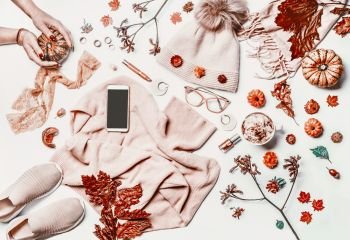 Autumn fashion woman clothes and shoes flat lay layout with smartphone mock up screen, sweater, knitted hat, scarf, eyeglasses, cosmetics,pumpkin,  cappuccino and fall leaves. Female hands.White desk