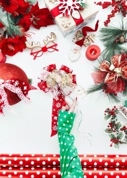 Female hand in green polka dot blouse making festive Christmas funky bow on white desktop with various red holiday decoration, ribbons and wrapping paper. Top view. Copy space. Flat lay