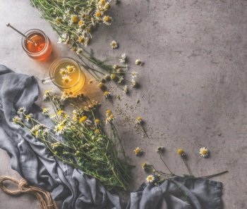 Fresh chamomile tea in glass cup, honey and honey spoon, medicinal plants. Natural treatment with herbs. Dark cloth and cord on dark concrete background. Top view.