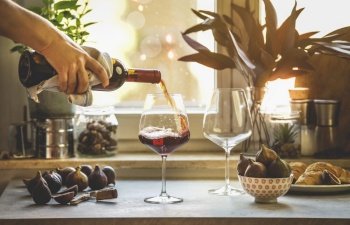 Hand pouring red wine from bottle in two wine glasses on kitchen table with figs, wine cutlery and cork at window background with plants and natural sunlight. Modern still life