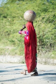 traditionally dressed woman in traditional indian Saree walking at rural road with pot at head. Rajasthan. India