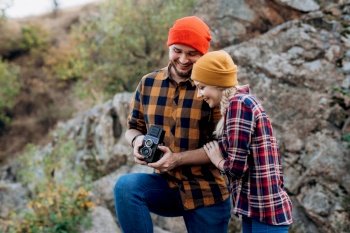 Bald guy with a beard and a blonde girl in bright hats on the background of the river are taking pictures with an old camera