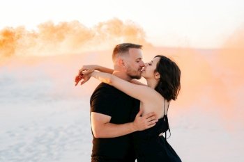 guy and a girl in black clothes hug and run on the white sand with orange smoke