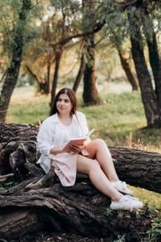 young girl in white clothes sits on a tree in the forest and reads a book