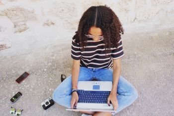 A portrait of focused young black woman with curly hair wearing glasses, jeans and a striped t-shirt, next to technology as smartphones and cameras, sitting on the ground and working or making homework