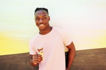 Young handsome black man wears a pink t-shirt, smiles and holds a ice cream cone in summertime on a painted wall as a sunrise or sunny day