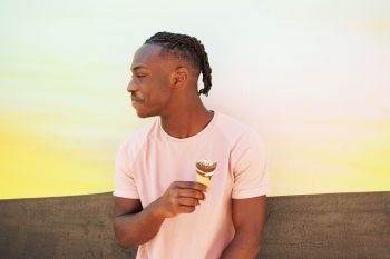 Young handsome black man wears a pink t-shirt, holds and eats a ice cream cone in summertime on a painted wall as a sunrise or sunny day
