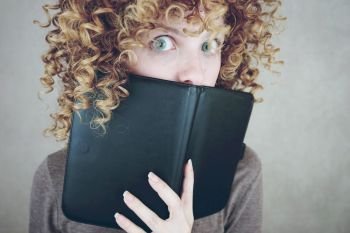 Close-up portrait of a beautiful and young funny woman with blue eyes and curly blonde hair, she is behind of a agenda or ebook and she is surprised