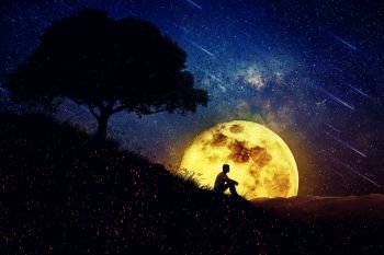 Boy sit alone on a hill in the center of nature, over a full moon night background. Standing away from the crowd, waiting for the healing power of the nature.
