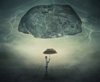 Surrealistic image as a man stand in a foggy street under a huge levitating rock, holding an umbrella in hand as a protection from danger. Life pressure and stress concept.