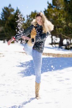 Young happy woman kicking snow in a snow-covered forest in the mountains in Sierra Nevada, Granada, Spain. Female wearing winter clothes playing with snow.. Young happy woman kicking snow in a snow-covered forest in the mountains