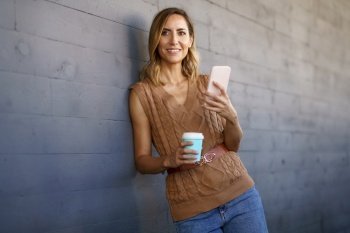 Woman in stylish knitted top, with cup of coffee to go looking away with smile and using smartphone while leaning on gray wall on city street. Stylish female with coffee browsing cellphone
