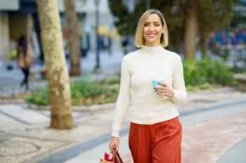 Positive female wearing turtleneck and pants walking with bag and holding cup of takeaway coffee drink in park. Smiling woman with cup of beverage carrying paper bag