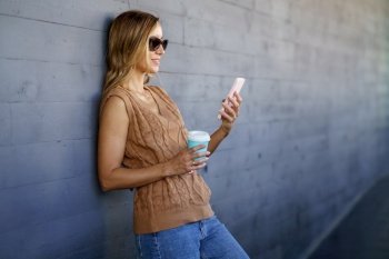 Glad female in stylish clothes and sunglasses, with cup of coffee smiling and browsing social media on cellphone while leaning on gray wall on city street. Woman with coffee leaning on wall and using smartphone