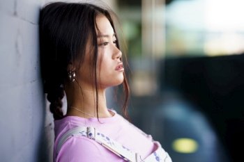 Young Asian female in casual clothes with braid leaning on wall and looking away on city street in daytime. Asian woman leaning on wall