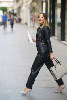 Side view of confident stylish woman in leather outfit and heels carrying modern handbag with eco friendly bottle and walking in city. Modern trendy female with reusable bottle in downtown