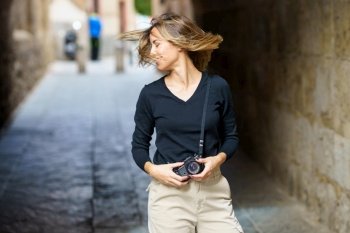 Positive young female traveler in casual clothes with vintage photo camera, smiling and shaking hair while standing on narrow path during sightseeing tour in old town. Happy female photographer smiling and shaking hair on street