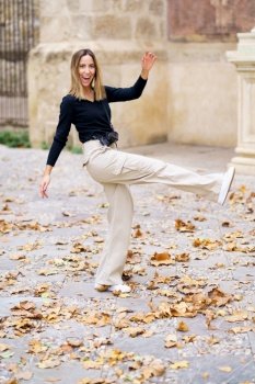 Side view of delighted young female tourist with long blond hair in casual clothes, smiling brightly and looking at camera while kicking golden fallen leaves during autumn holidays. Cheerful young woman kicking fallen leaves while walking in park