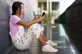 Full body young Asian woman in stylish clothes with cup of coffee to go siting on slope outside modern building and browsing smartphone in daytime. Asian female with coffee using cellphone on slope