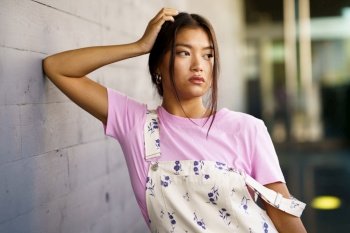 Young Asian woman in pink t shirt and stylish overalls touching head and looking away while leaning on gray wall on city street. Stylish Asian female leaning on wall