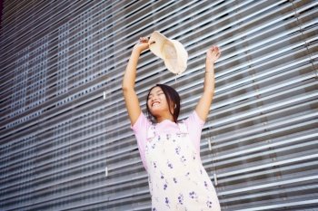 From below happy young Asian female in stylish clothes, smiling with closed eyes and raising arms with cap while dancing against metal wall on city street. Young Asian woman dancing with raised arms
