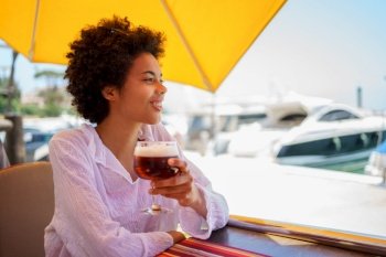 Cheerful young African American female in white blouse with curly hair smiling and looking away, while enjoying beer in street restaurant on sunny summer day on embankment. Happy black woman resting in street restaurant