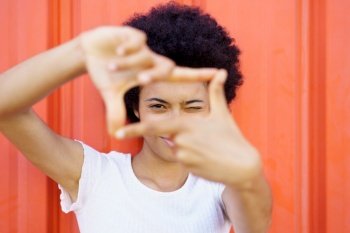 Glad African American female in casual wear winking and looking at camera through frame gesture on red background in city. Content black woman making frame gesture near wall