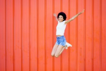 Full body of smiling African American female in casual outfit looking at camera while jumping with outstretched arms and bent knees against red background. Carefree black woman jumping near wall
