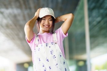 Merry young Asian woman in stylish t shirt and overalls with cap, looking at camera with smile and touching head on blurred background of contemporary building in city. Cheerful Asian female touching head