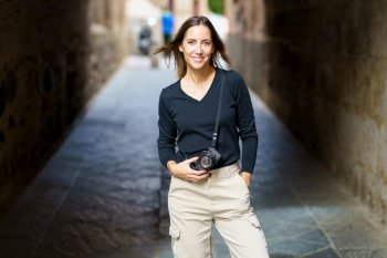 Self assured young female photographer with camera in casual clothes standing on narrow street with hand in pocket and looking at camera during vacation. Smiling woman with photo camera looking at camera on narrow street