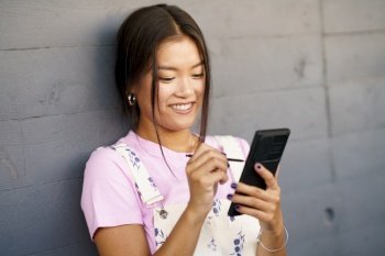 Happy young Asian female in stylish clothes smiling and using stylus to browse social media on cellphone while leaning on gray wall on city street. Cheerful Asian woman using smartphone near wall