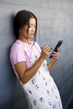 Young Asian woman in stylish clothes browsing news on mobile phone with stylus while leaning on gray wall on city street. Asian female leaning on wall and using cellphone