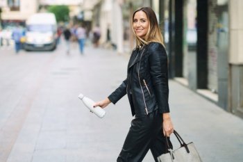 Side view of stylish female in leather pants and jacket carrying bag and takeaway reusable bottle while commuting on town street looking at camera. Smart casual lady with bag and bottle walking on street