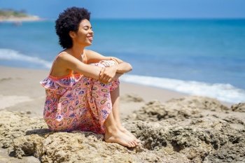 Cheerful young black female traveler in summer dress, with curly hair embracing knees and smiling with closed eyes while sitting on uneven stones against waving sea on sunny day on beach. Happy African American woman spending time on beach