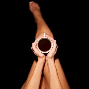 Top view of crop unrecognizable barefooted female sitting in black studio with cup of hot coffee. Crop anonymous woman with coffee cup in black studio