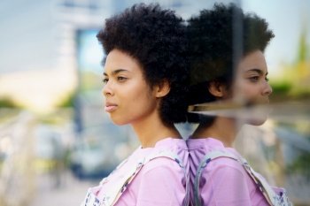 Side view of serious African American female with Afro hairstyle standing near glass house on street against blurred background in city. Black woman near glass building in city